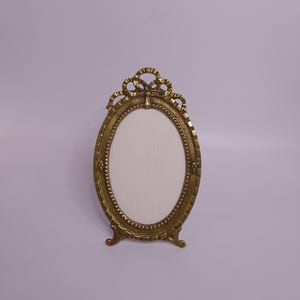 Può includere: Una cornice ovale in stile antico, color oro. La cornice ha un fiocco decorativo nella parte superiore e un bordo perlato. Poggia su due piedini. Il centro è un ovale bianco.
