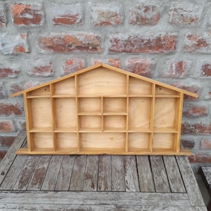 Può includere: Una mensola espositiva in legno a forma di casa, con numerosi piccoli scomparti. La mensola in legno chiaro è progettata per esporre piccoli oggetti. È appoggiata a un muro di mattoni e a un tavolo di legno.