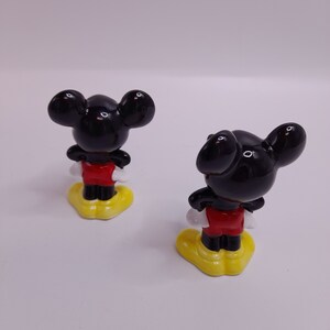 May include: Two ceramic Mickey Mouse figurines, both facing away from the camera. The figurines are black with red shorts and yellow shoes.