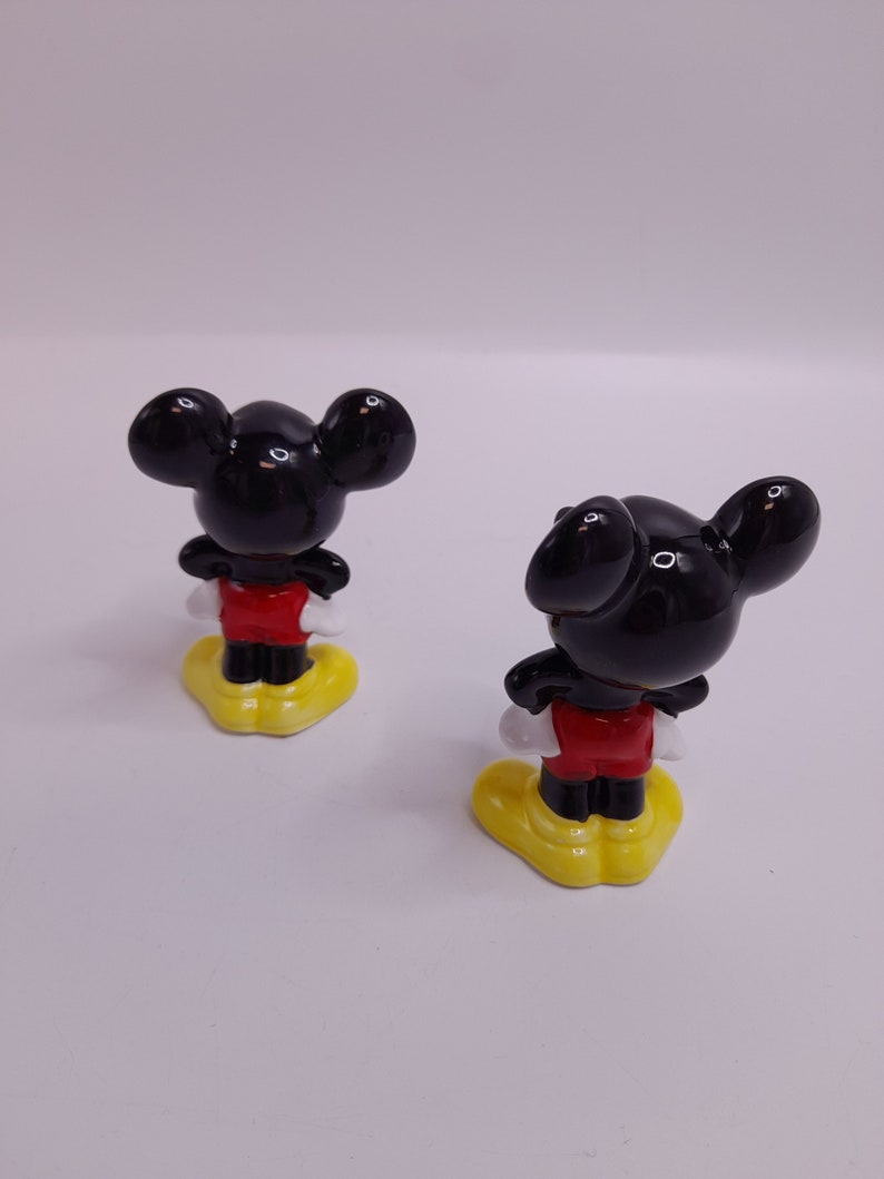May include: Two ceramic Mickey Mouse figurines, both facing away from the camera. The figurines are black with red shorts and yellow shoes.