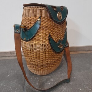May include: A woven wicker bag with a brown leather strap and green leather accents. The bag has a brass clasp and a small brass bird design on the front.