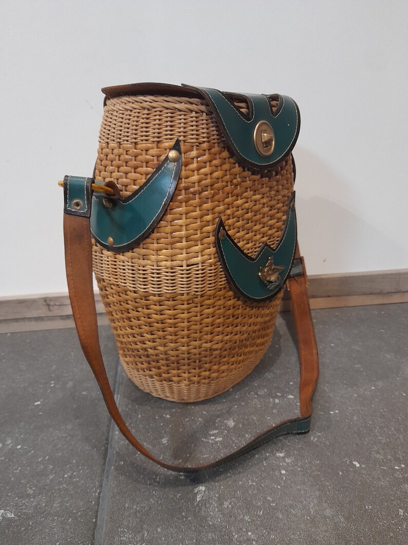 May include: A woven wicker bag with a brown leather strap and green leather accents. The bag has a brass clasp and a small brass bird design on the front.