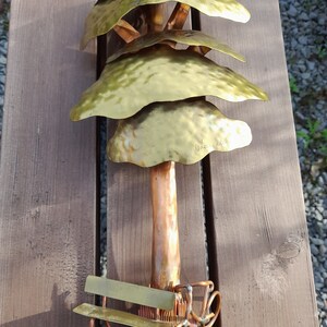 May include: A gold metal sculpture of a tree with a bench and a bicycle underneath. The tree has four layers of leaves and the trunk is copper coloured. The sculpture is signed "Dhavala".