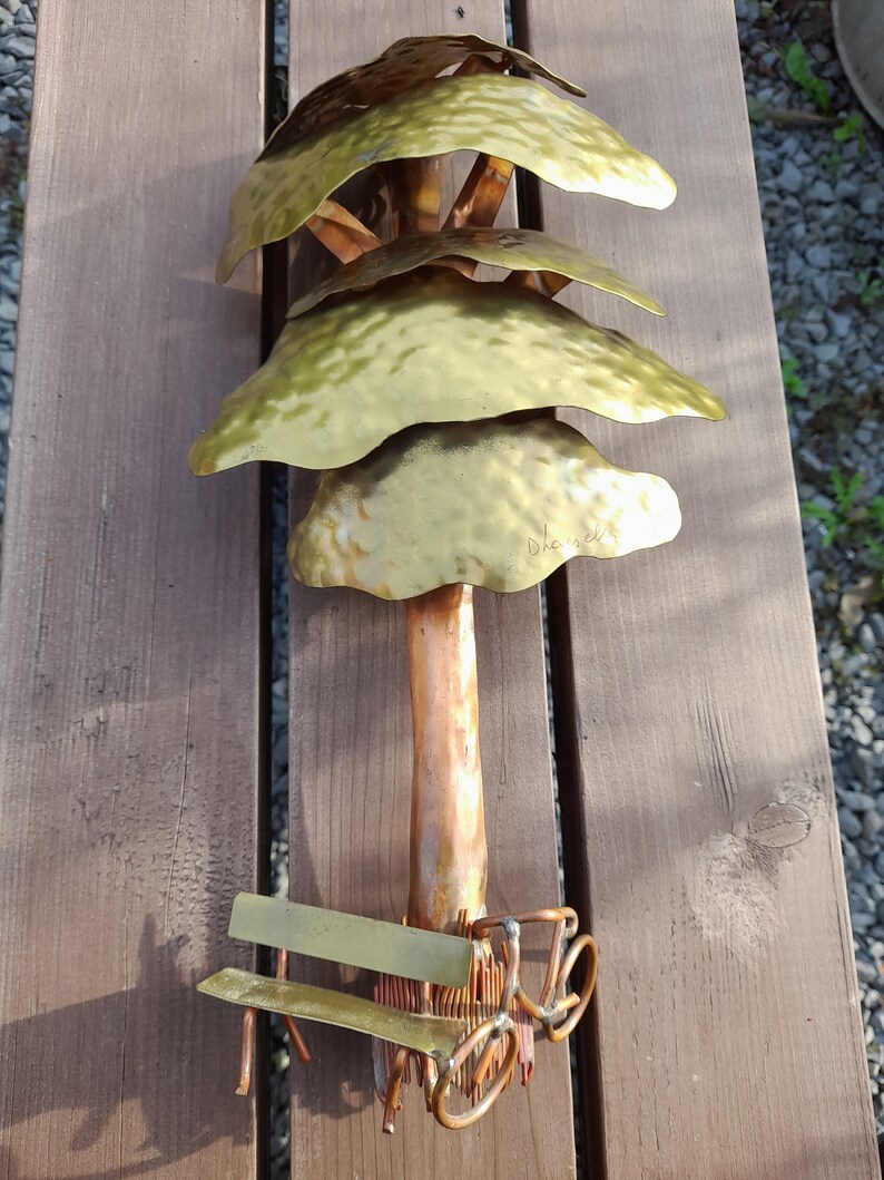 May include: A gold metal sculpture of a tree with a bench and a bicycle underneath. The tree has four layers of leaves and the trunk is copper coloured. The sculpture is signed "Dhavala".
