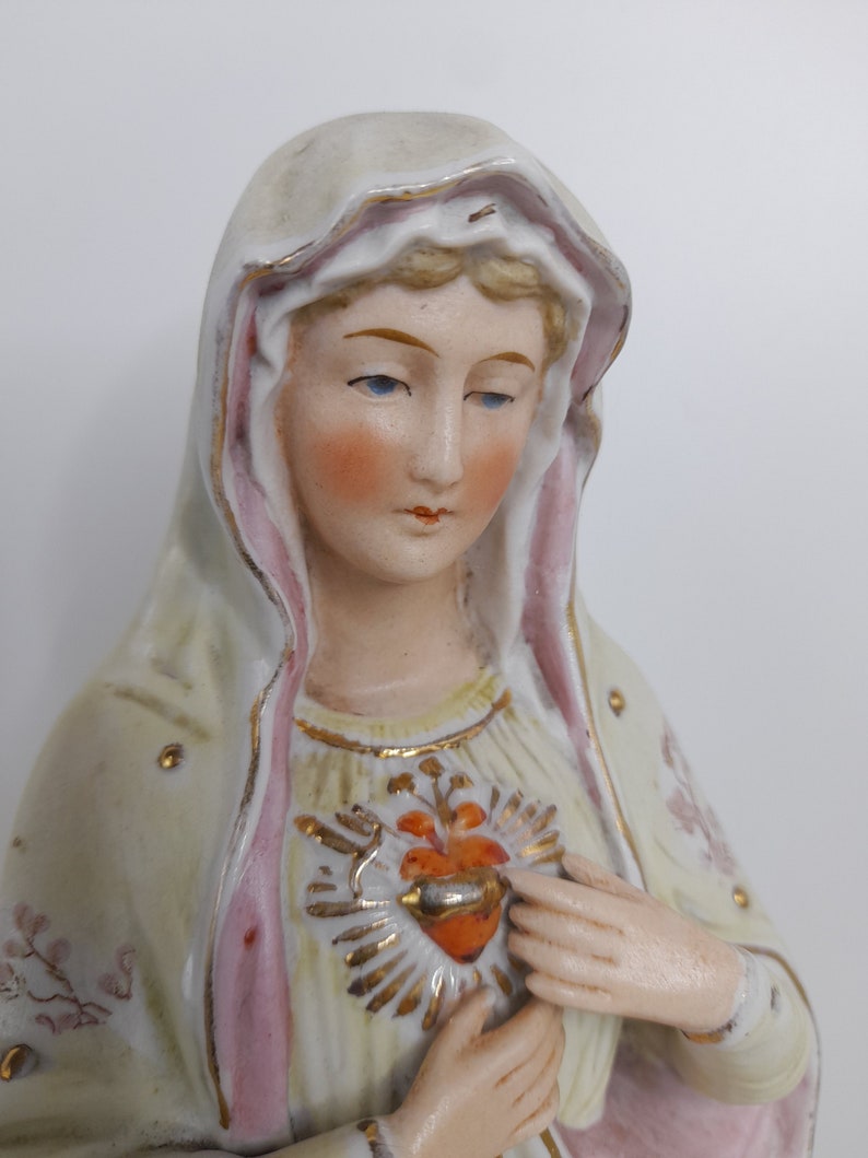 Puede incluir: Una figurita de porcelana de la Virgen Mar&iacute;a, vestida con una t&uacute;nica blanca y rosa con detalles dorados. Tiene un coraz&oacute;n con una corona de espinas en el pecho.