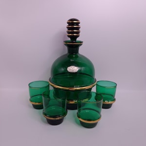 May include: A vintage green glass decanter set with a round decanter and six matching shot glasses. The decanter has a gold rim and a black and gold stopper. The shot glasses have gold rims and black bases. The set is likely for serving liquor.