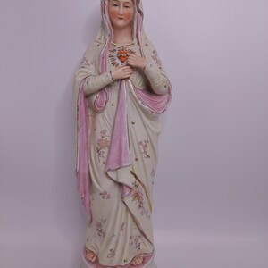 Puede incluir: Una estatua de porcelana de la Virgen Mar&iacute;a con una t&uacute;nica rosa y blanca, sosteniendo un coraz&oacute;n en sus manos. La estatua est&aacute; sobre una base con dise&ntilde;os florales.