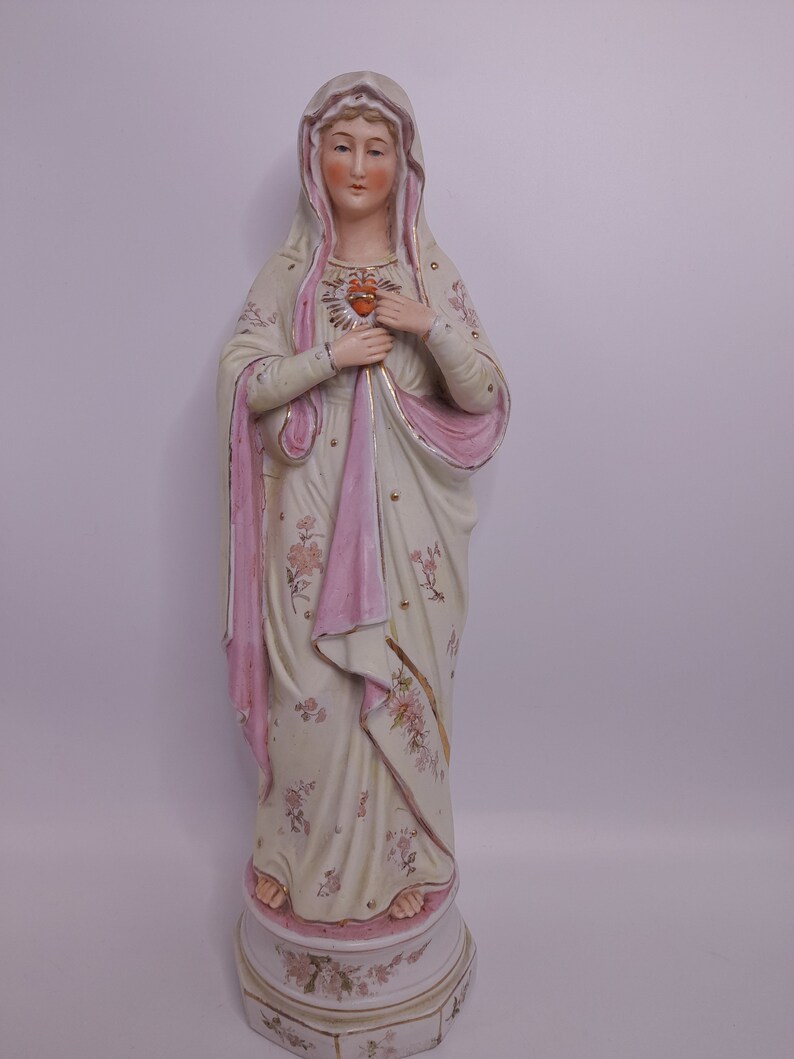 Puede incluir: Una estatua de porcelana de la Virgen Mar&iacute;a con una t&uacute;nica rosa y blanca, sosteniendo un coraz&oacute;n en sus manos. La estatua est&aacute; sobre una base con dise&ntilde;os florales.