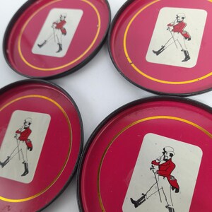 Op de afbeelding: Set van zes rode metalen onderzetters met een gouden rand en zwarte randen. Elke onderzetter heeft een witte rechthoek met een illustratie van een heer in rode kleding. De heer draagt een hoed en houdt een wandelstok vast.