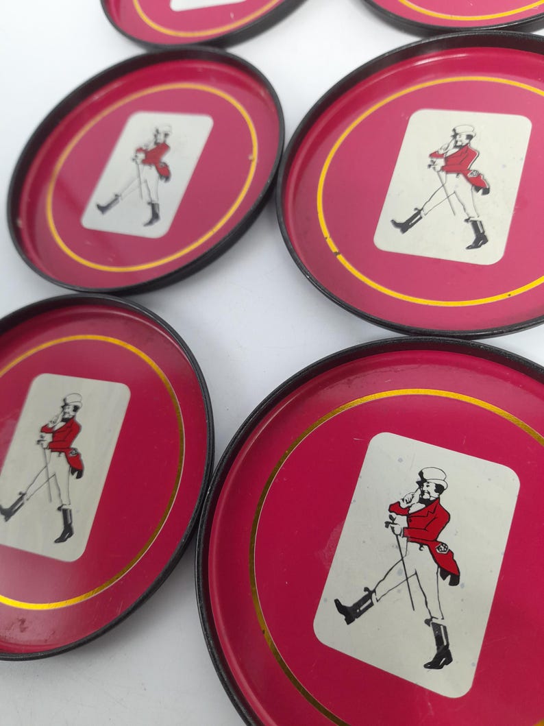 Op de afbeelding: Set van zes rode metalen onderzetters met een gouden rand en zwarte randen. Elke onderzetter heeft een witte rechthoek met een illustratie van een heer in rode kleding. De heer draagt een hoed en houdt een wandelstok vast.