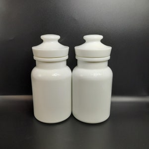 以下が含まれることがあります： お揃いの蓋が付いた白いセラミックジャー2個。ジャーは円筒形の本体と、小さな円形の蓋が付いた丸みを帯びた上部を備えています。背景は無地の黒です。
