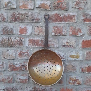 May include: A round, metallic colander with a warm, burnished copper-toned finish and a long, flat handle featuring a circular hanging loop at the top. The bowl of the colander is perforated with numerous small, evenly spaced circular drainage holes.
