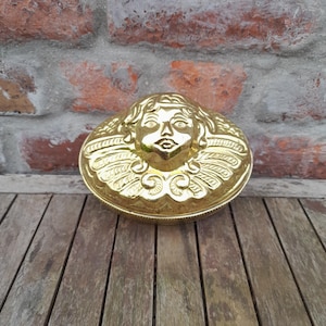 May include: A shiny gold-colored metal mold featuring a raised relief of a cherub face with wings. The oval-shaped object has a smooth, reflective surface and detailed embossed features depicting the facial structure and feathered wing patterns.