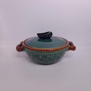 May include: A ceramic soup tureen with a green basket weave design. The tureen has a matching green lid with a black handle. The rim is trimmed with a brown border, and it has brown handles on either side.