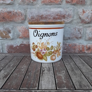 May include: A white ceramic kitchen storage jar, labelled "Oignons" in black script. It has a decorative floral and fruit design in brown, orange, and green. The jar has a wooden lid and a brown rim, suitable for storing onions.