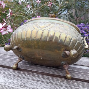 Pu&ograve; includere: Un vaso di fiori vintage in ottone con dettagli ornati. Il vaso ha un corpo arrotondato, tre piedini e manici decorativi. L'ottone ha una calda patina invecchiata. Lo sfondo presenta fiori e fogliame.
