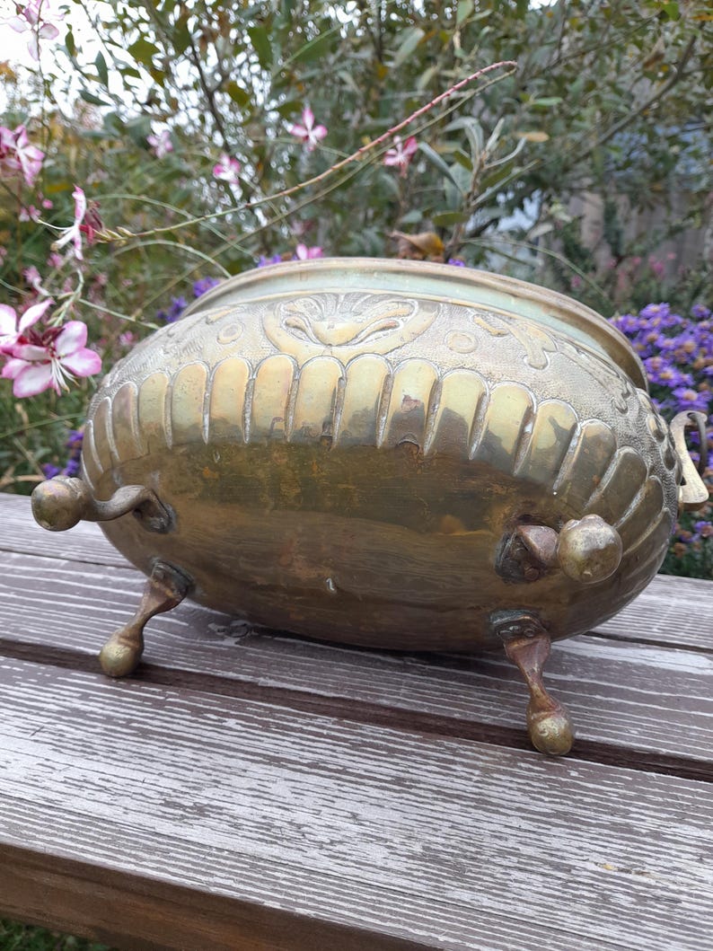 Pu&ograve; includere: Un vaso di fiori vintage in ottone con dettagli ornati. Il vaso ha un corpo arrotondato, tre piedini e manici decorativi. L'ottone ha una calda patina invecchiata. Lo sfondo presenta fiori e fogliame.