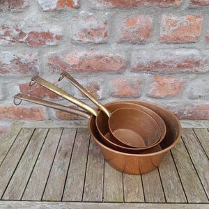 May include: Three copper pots and pans with brass handles are nested together on a wooden surface. The pots have a shiny, reflective surface. The background features a weathered brick wall.