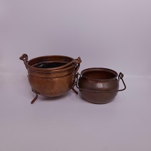 Könnte beinhalten: Zwei Kupfertöpfe mit Griffen. Der größere Topf hat drei Beine und ist leicht angelaufen. Der kleinere Topf ist glänzend und hat einen einzigen Griff.
