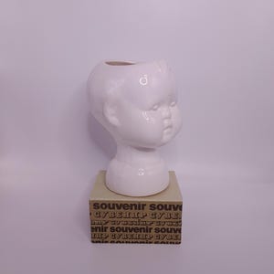 Può includere: Un vaso a forma di testa di bambino in ceramica bianca. Il vaso è su una piccola scatola rettangolare con la parola "souvenir" stampata ripetutamente in un carattere marrone. Il vaso ha una finitura liscia e lucida.