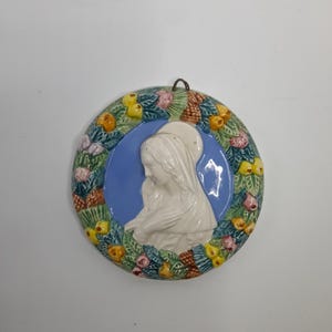 Può includere: Un'applique da parete rotonda in ceramica con un rilievo bianco di una figura religiosa su sfondo blu. La figura è circondata da una ghirlanda di foglie verdi, fiori gialli e arancioni e dettagli rosa. Un anello di metallo è attaccato per appenderla.