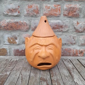 May include: A terracotta-colored ceramic pot with a face design. The pot has a conical lid with a hole at the top. The face has a furrowed brow, a downturned mouth, and small ears. The pot is sitting on a wooden surface.