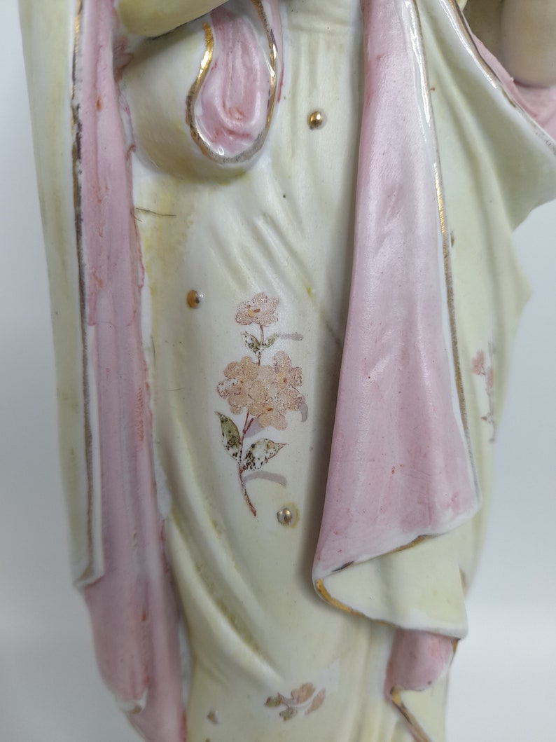 Puede incluir: Una figurita de porcelana de una mujer vestida con un vestido blanco con detalles rosas y florales. El vestido tiene un dise&ntilde;o fluido y la mujer sostiene un objeto peque&ntilde;o en su mano.