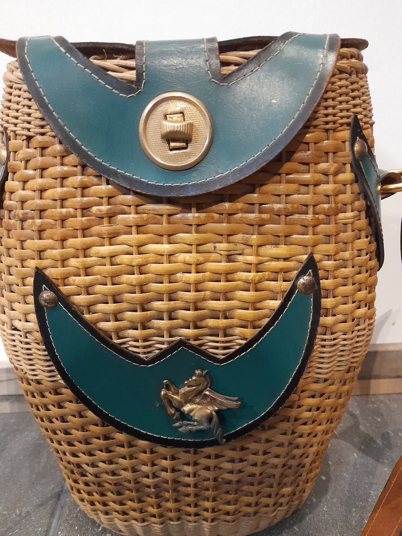 May include: A woven wicker basket with a teal leather flap and a metal button closure. The basket has a teal leather patch with a metal horse design.