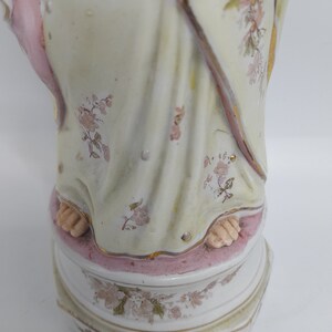 Puede incluir: Una figurita de porcelana de una mujer con un vestido blanco con detalles en rosa y dorado. El vestido est&aacute; decorado con motivos florales. La figurita est&aacute; de pie sobre una base con un borde rosa y motivos florales.