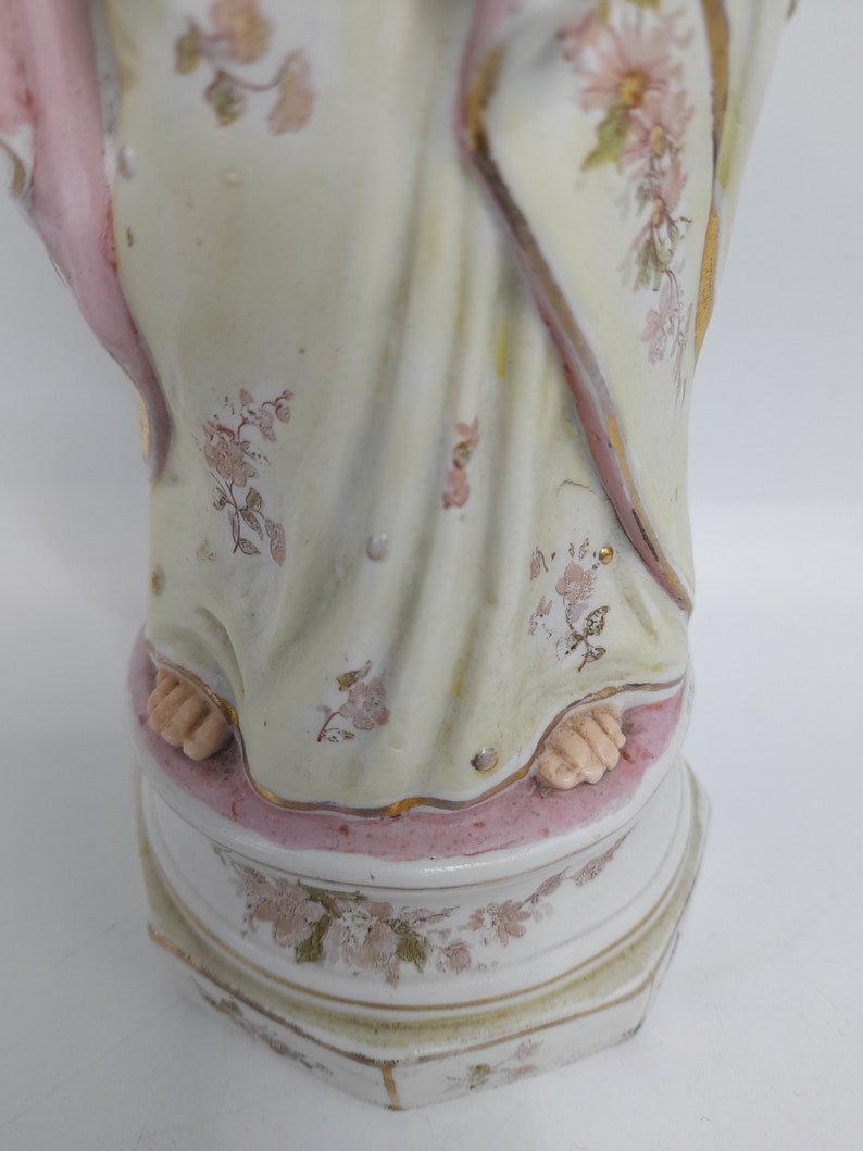 Puede incluir: Una figurita de porcelana de una mujer con un vestido blanco con detalles en rosa y dorado. El vestido est&aacute; decorado con motivos florales. La figurita est&aacute; de pie sobre una base con un borde rosa y motivos florales.