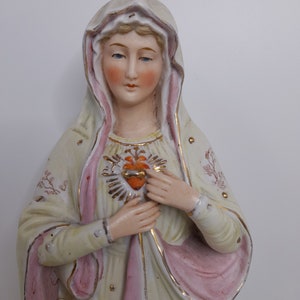 Puede incluir: Una figurita de porcelana de la Virgen Mar&iacute;a, vestida con una t&uacute;nica blanca y rosa con detalles dorados. Ella sostiene un coraz&oacute;n con un centro rojo y un borde dorado.