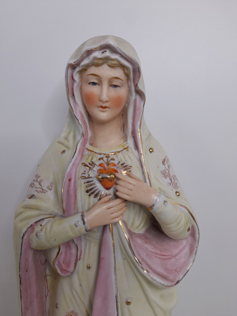 Puede incluir: Una figurita de porcelana de la Virgen Mar&iacute;a, vestida con una t&uacute;nica blanca y rosa con detalles dorados. Ella sostiene un coraz&oacute;n con un centro rojo y un borde dorado.