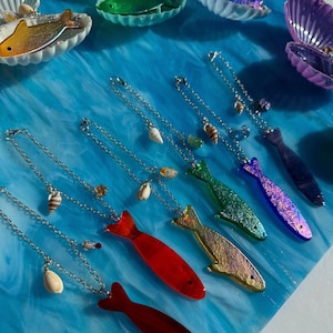 May include: A collection of fish-shaped pendants in various colors, including red, gold, green, blue, and purple, with silver chains and seashell accents. The pendants are displayed on a blue, textured surface, alongside seashell-shaped containers.