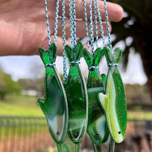 May include: A group of green glass fish ornaments with white accents hanging from white and blue strings. The fish have a swirl pattern and are suspended in mid-air.