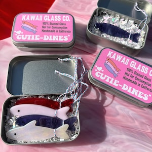以下が含まれることがあります： ピンク色のラベルと魚の形をしたガラスの装飾品が付いた、いくつかの小さな銀色の金属製の缶。ラベルには「KAWAII GLASS CO.」と「CUTIE-DINES」と書かれています。装飾品は赤、ピンク、青、白など様々な色があります。缶には装飾用の紙が詰められています。