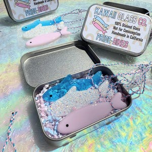 May include: Fish-shaped necklaces in a metal tin. The necklaces are blue, white, and pink, with a braided cord. The tin has the text "KAWAII GLASS CO." and "PRIDE-DINES". The necklaces are made of 100% stained glass and handmade in California.