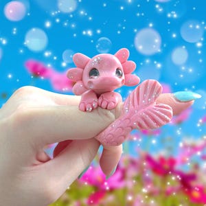 May include: A pink axolotl ring featuring a sculpted creature with large dark eyes, external gills, and a textured tail that wraps around the finger. The surface of the pink figure is accented with silver glitter throughout its body.
