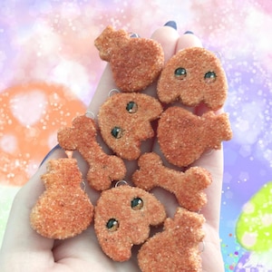 May include: A selection of orange, textured charms in diverse forms, such as hearts, bone shapes, and rounded figures with small, dark eyes. The charms are held in a hand against a soft, colourful background.