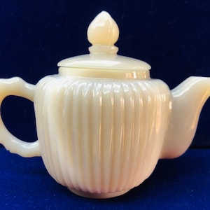 May include: A small, white, ceramic teapot with a ribbed design and a lid. The teapot has a handle and a spout.