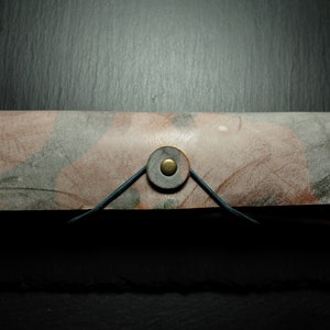 Könnte beinhalten: Ein braunes Leder-Bleistift-Etui mit einem grauen und rosafarbenen Marmormuster. Das Etui hat einen Messingknopfverschluss und einen schwarzen Lederriemen. Die Initialen "HC" sind auf der rechten Seite des Etuis geprägt.
