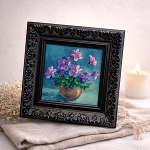 Op de afbeelding: Een ingelijst schilderij van paarse en roze bloemen in een bruine pot tegen een blauwe achtergrond. Het kunstwerk is gevat in een zwart, sierlijk frame. Het schilderij rust op een neutraal gekleurde doek.