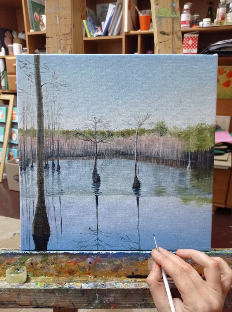 Puede incluir: Una pintura sobre lienzo que representa una escena de pantano con &aacute;rboles altos y delgados reflejados en el agua. El cielo es de un azul claro y el agua es de un azul verdoso claro. Los &aacute;rboles son de color marr&oacute;n oscuro y el agua es de un azul verdoso claro.