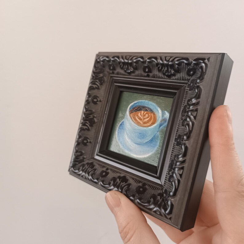 Puede incluir: Una peque&ntilde;a pintura enmarcada de una taza de caf&eacute; azul con un dise&ntilde;o de arte latte marr&oacute;n. El marco es negro con detalles ornamentados.
