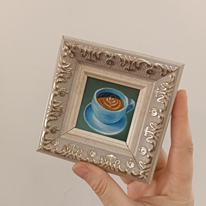 Puede incluir: Una peque&ntilde;a pintura enmarcada de una taza de caf&eacute; azul con un dise&ntilde;o de arte latte. La taza est&aacute; sobre un platillo azul y el fondo es verde.