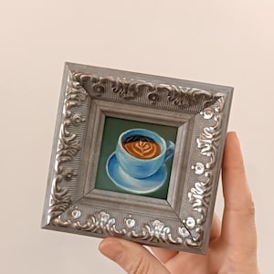 Puede incluir: Una peque&ntilde;a pintura enmarcada de una taza de caf&eacute; azul con un dise&ntilde;o de arte latte. El marco es plateado con un dise&ntilde;o ornamentado.
