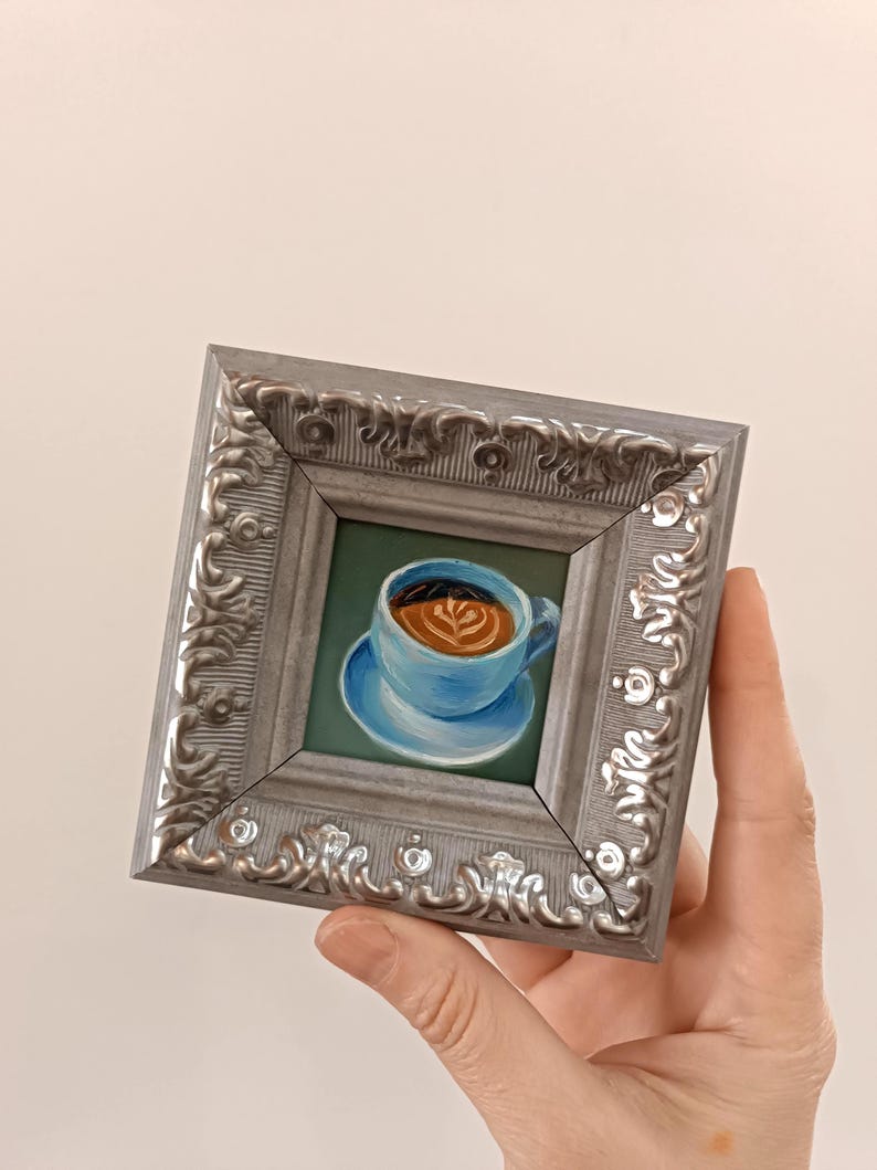 Puede incluir: Una peque&ntilde;a pintura enmarcada de una taza de caf&eacute; azul con un dise&ntilde;o de arte latte. El marco es plateado con un dise&ntilde;o ornamentado.