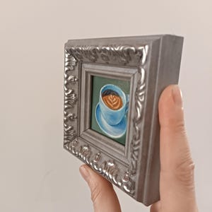 Puede incluir: Una peque&ntilde;a pintura enmarcada de una taza de caf&eacute; azul con un dise&ntilde;o de arte latte en la parte superior. La taza est&aacute; en un platillo y el fondo es de color verde.