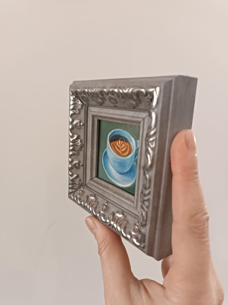 Puede incluir: Una peque&ntilde;a pintura enmarcada de una taza de caf&eacute; azul con un dise&ntilde;o de arte latte en la parte superior. La taza est&aacute; en un platillo y el fondo es de color verde.
