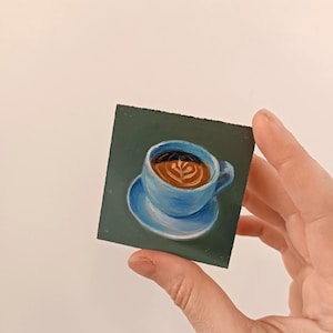 Puede incluir: Un primer plano de una peque&ntilde;a pintura de una taza de caf&eacute; azul con caf&eacute; marr&oacute;n y arte latte sobre un fondo verde.