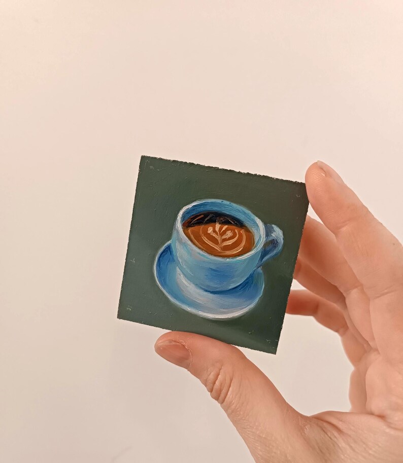 Puede incluir: Un primer plano de una peque&ntilde;a pintura de una taza de caf&eacute; azul con caf&eacute; marr&oacute;n y arte latte sobre un fondo verde.